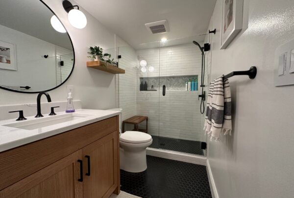 Remodeled bathroom with an oak vanity, matte black fixtures, white subway tile walk-in shower with glass door, and black hexagon floor tile