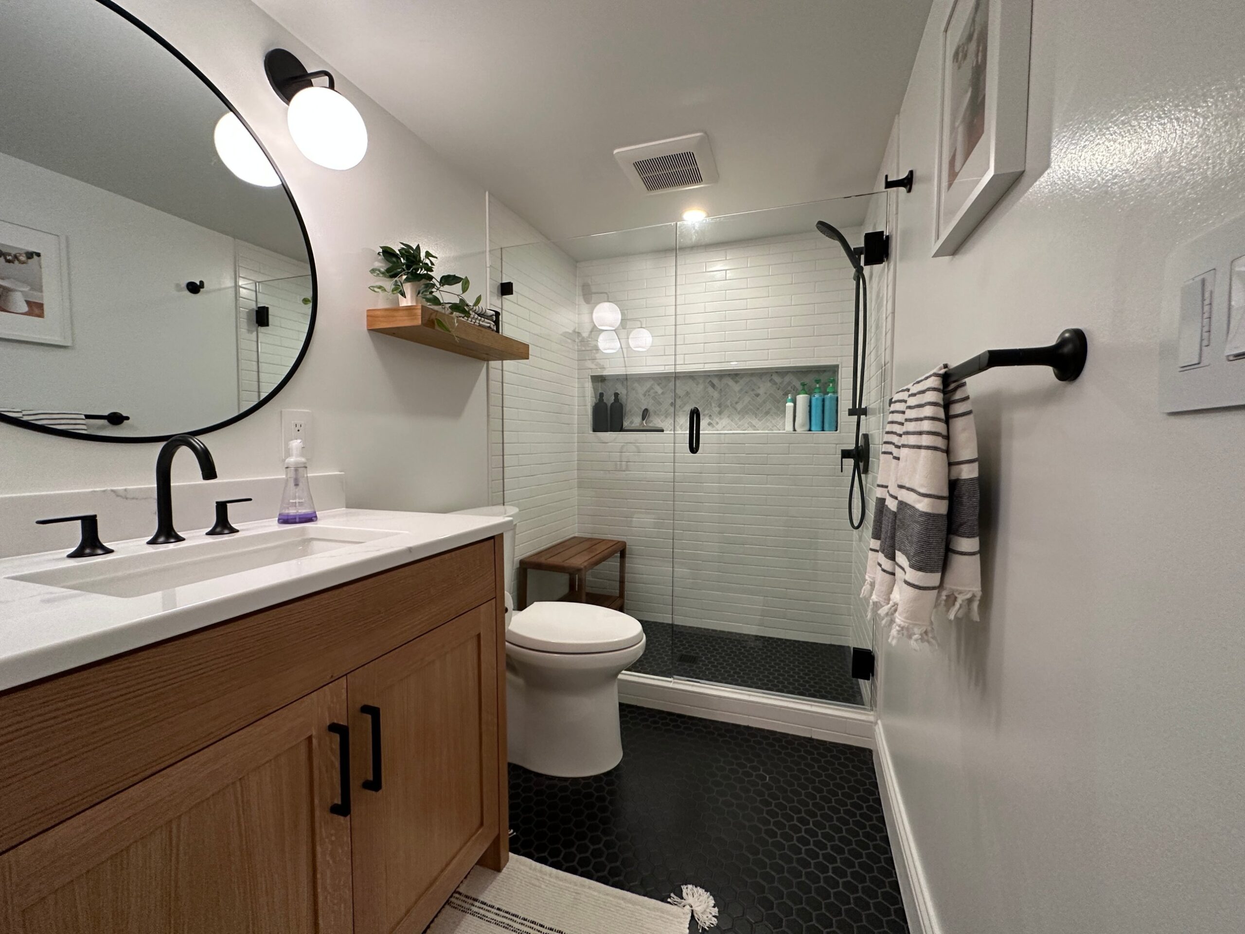Remodeled bathroom with an oak vanity, matte black fixtures, white subway tile walk-in shower with glass door, and black hexagon floor tile