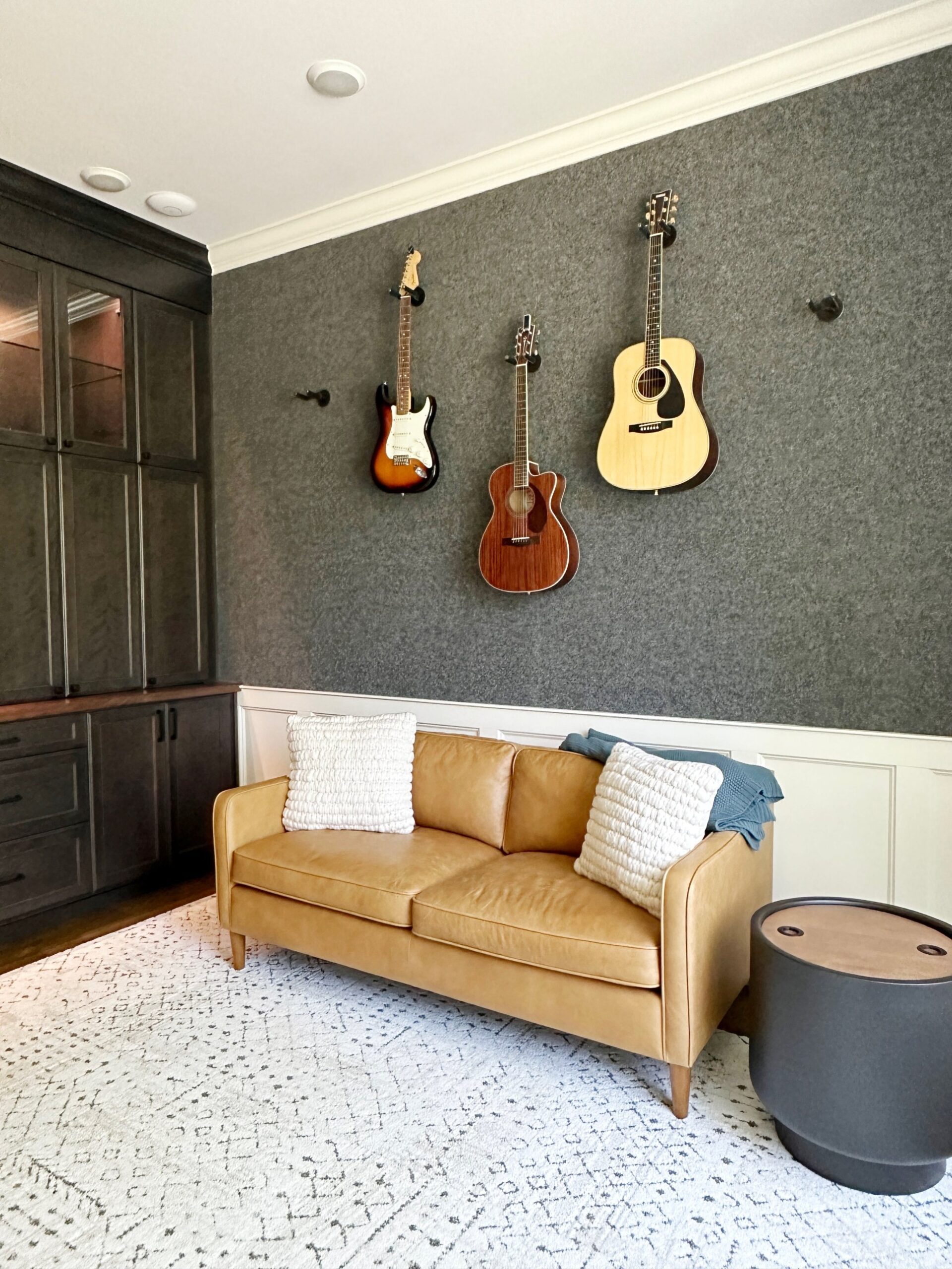 Music room with dark stained built-in cabinets, white wainscoting, a textured gray wall with three guitars, and a tan leather sofa