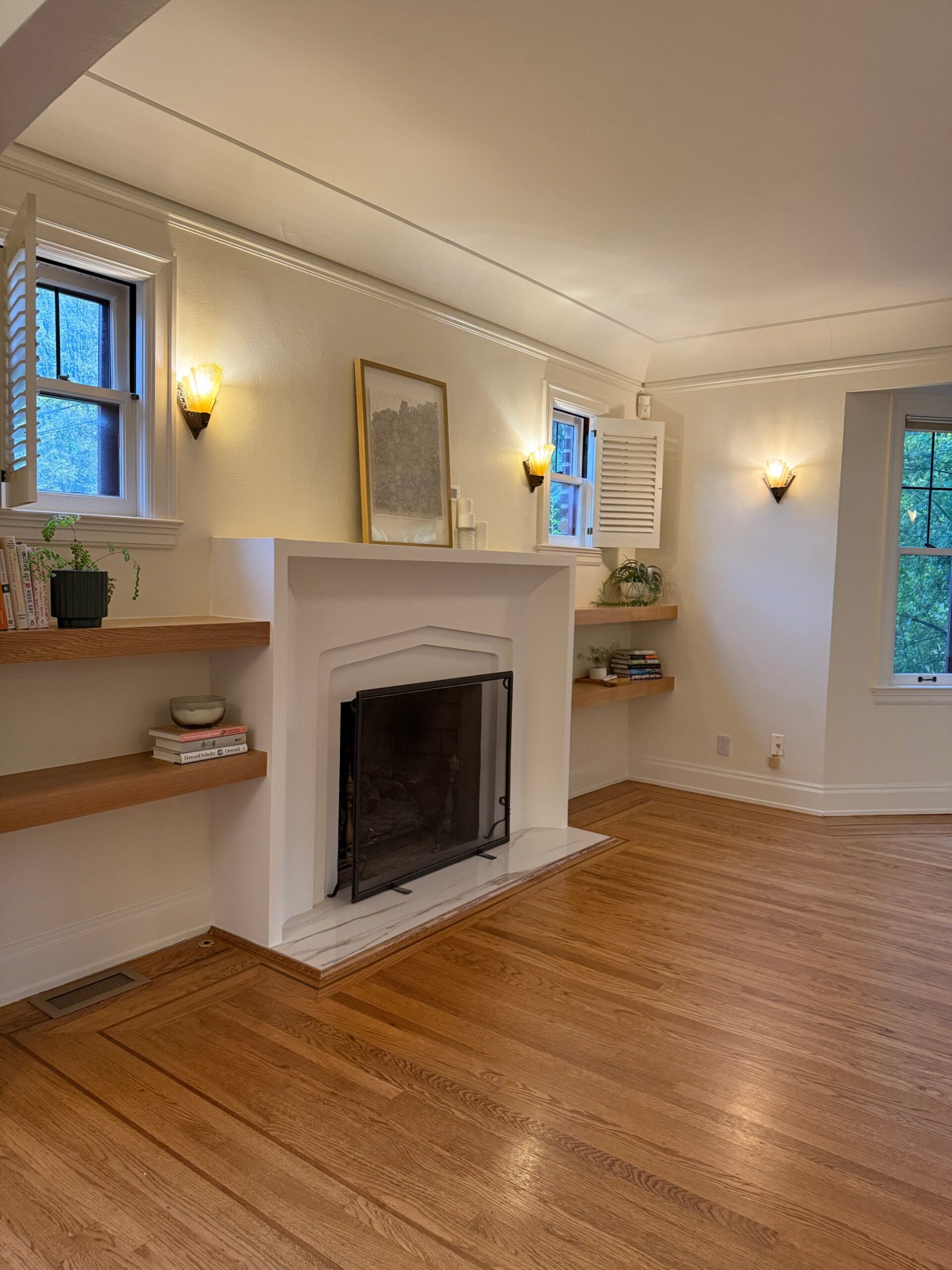 Living room in an older Seattle home after a remodel, with a white arched fireplace surround, oak floating shelves, wall sconces and refinished oak floors with an inlaid border