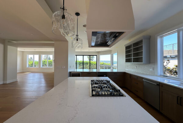 Open kitchen in a whole-home remodel, with a quartz island cooktop under a ceiling hood, glass pendant lights, and windows looking out to the water