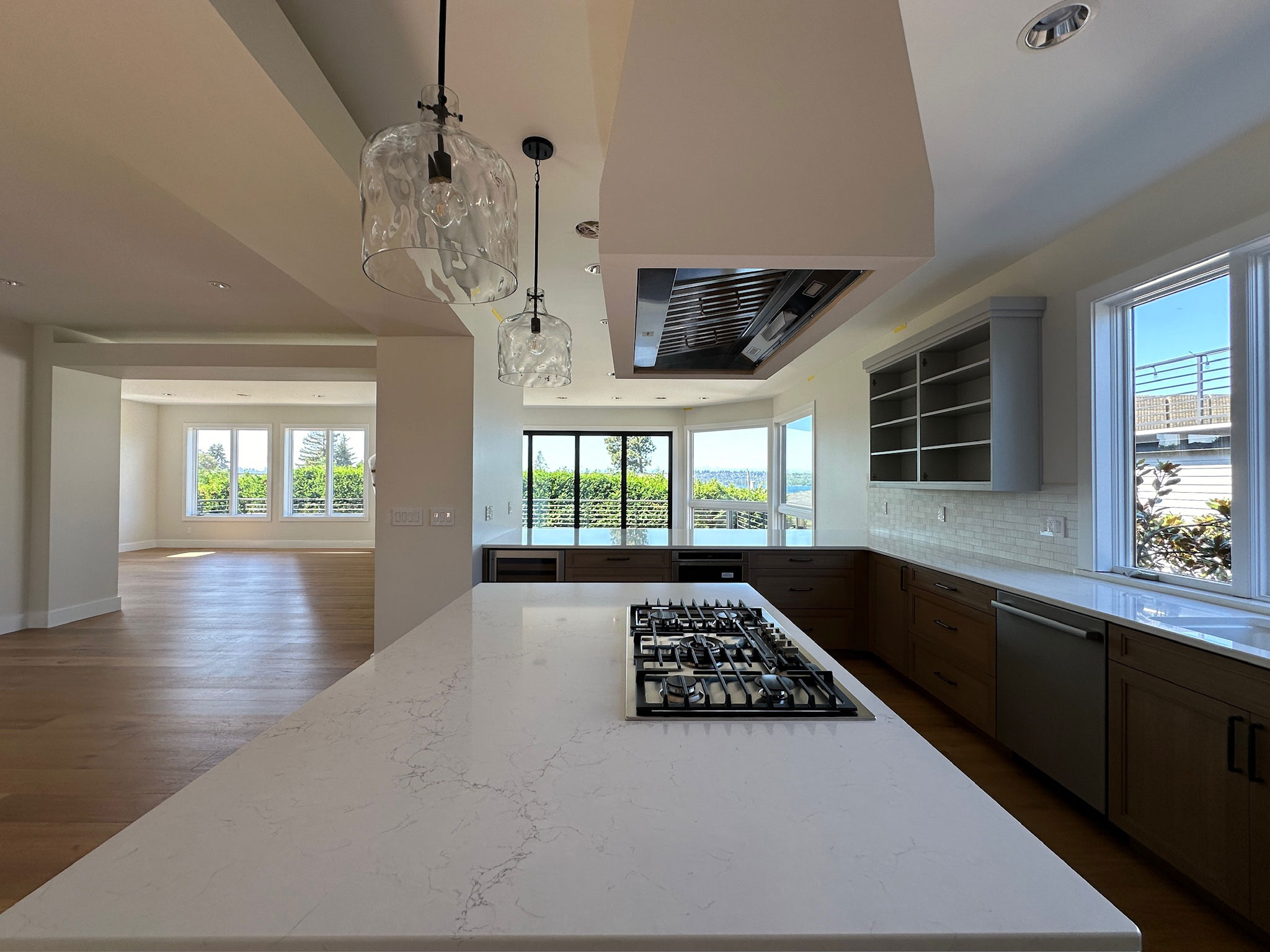 Open kitchen in a whole-home remodel, with a quartz island cooktop under a ceiling hood, glass pendant lights, and windows looking out to the water